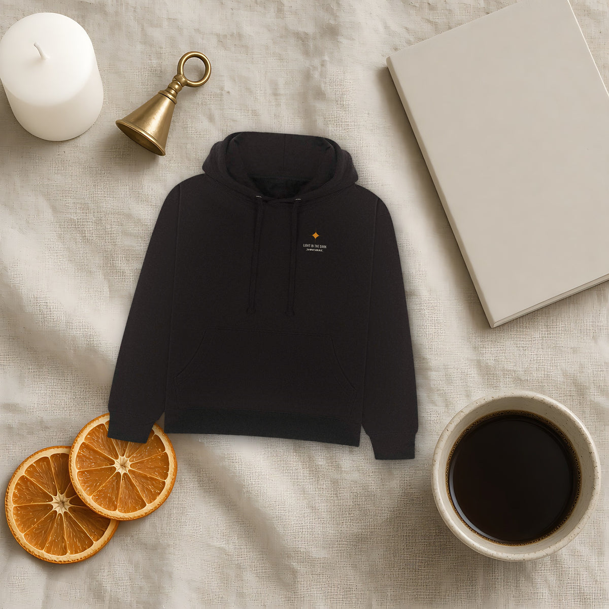 Black Light In The Dark Graphic Hoodie flat lay on linen with a candle, a brass bell, dried orange slices and coffee