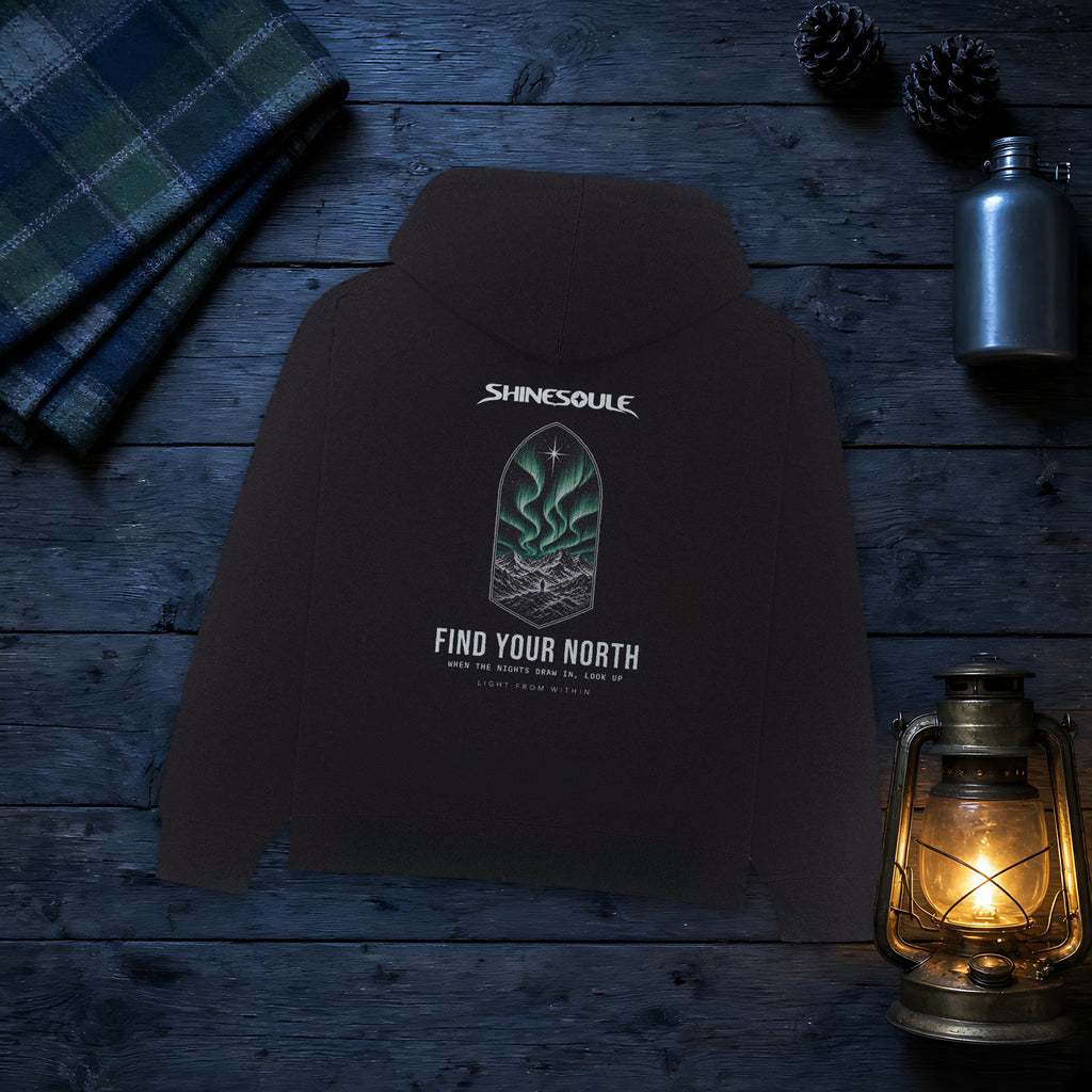 Black Northern Light Graphic Hoodie laid on dark wooden boards beside a tartan blanket, a flask and a lit lantern