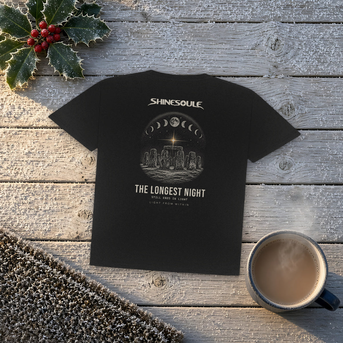 Black Solstice Graphic T-Shirt on weathered wooden boards with holly, a knitted throw and a cup of coffee