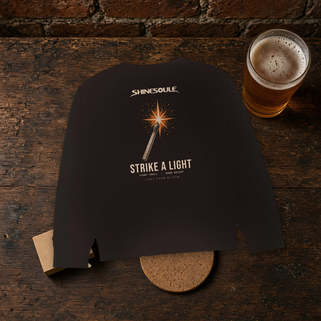 Black Strike A Light Graphic Crewneck Sweatshirt on a dark wooden bar beside a pint and a cork coaster