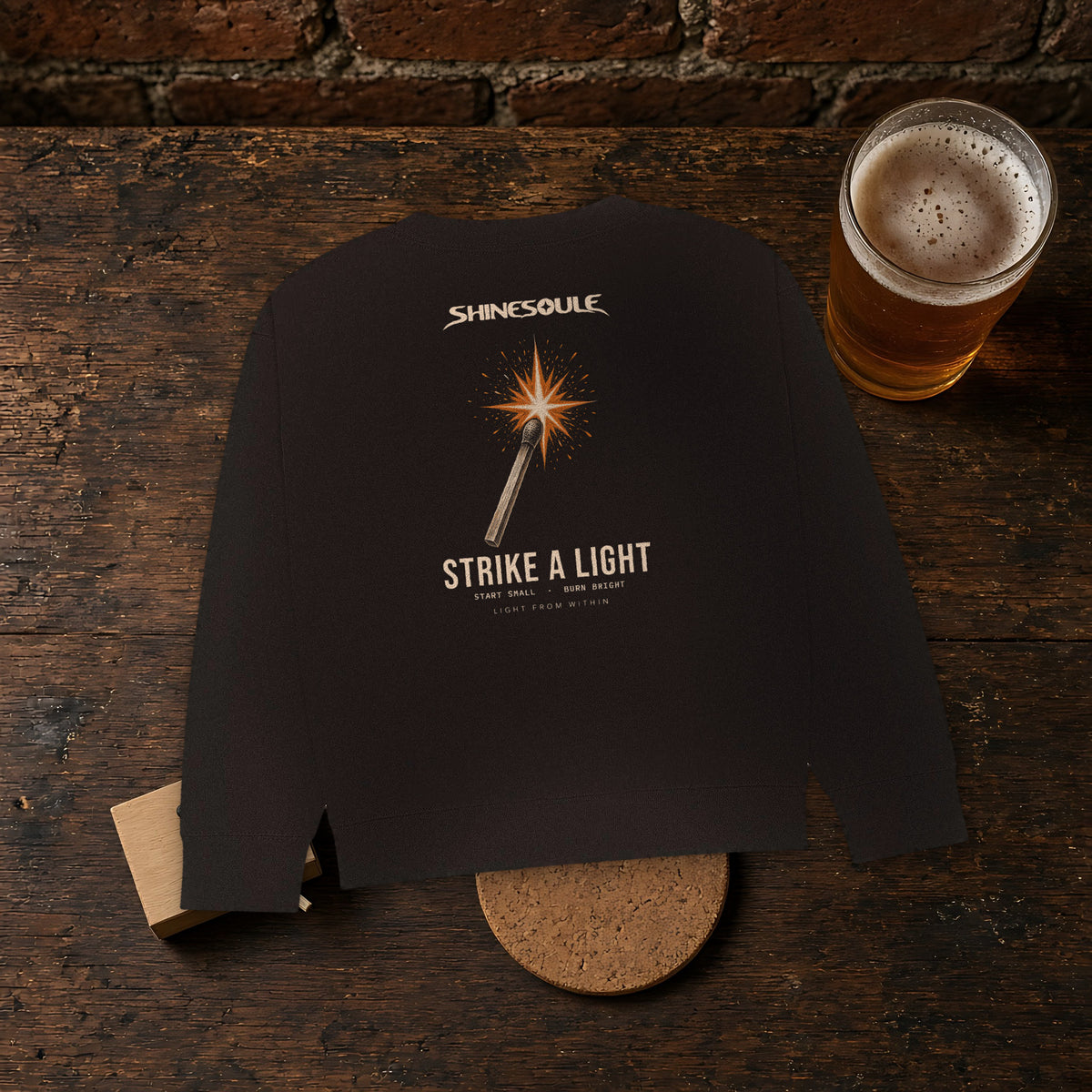 Black Strike A Light Graphic Crewneck Sweatshirt on a dark wooden bar beside a pint and a cork coaster